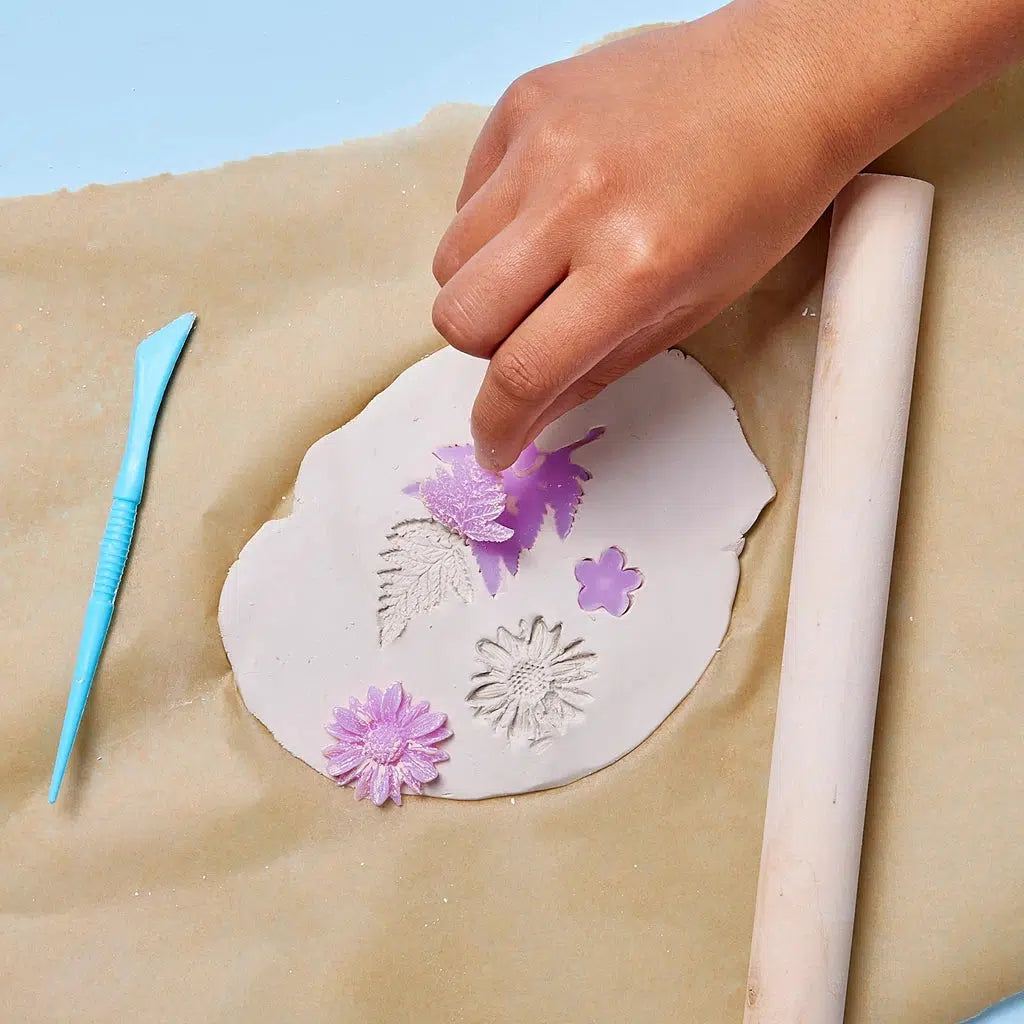 A hand arranges botanical stamps on clay, surrounded by a rolling pin and sculpting tool on parchment paper, setting the stage for delicate ceramic jewelry.