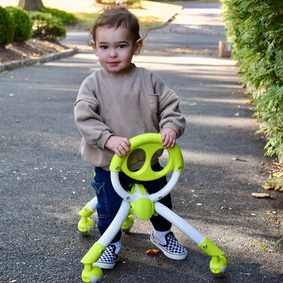 A child enhances motor skills with the Pewi Elite, a green and white developmental toy, as they navigate a paved path in a park surrounded by lush greenery.